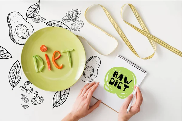 Top view of woman holding notebook with raw diet words near measuring tape and plate with diet lettering from vegetable slices on white background with leaves and avocado illustration — Stock Photo