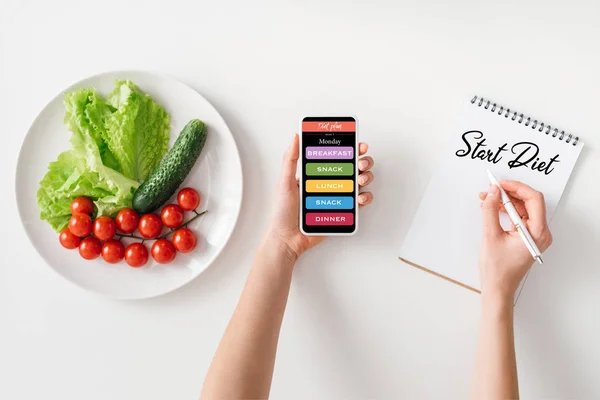Vue du dessus de la femme tenant smartphone et l'écriture dans un carnet avec commencer le régime alimentaire lettrage près des légumes mûrs sur plaque sur fond blanc — Stock Photo