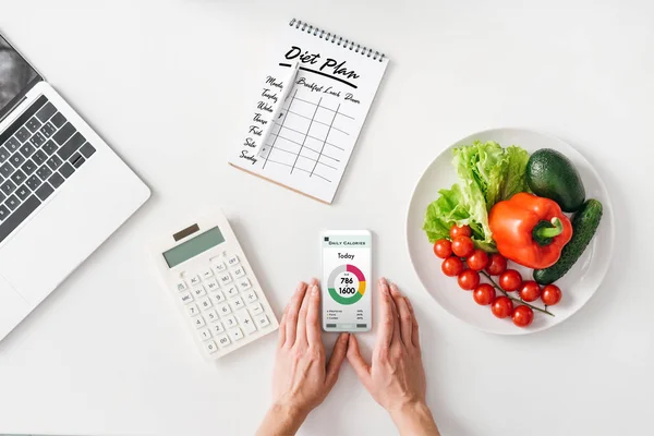 Visão superior da mulher segurando smartphone perto de calculadora, notebook e legumes no fundo branco — Fotografia de Stock