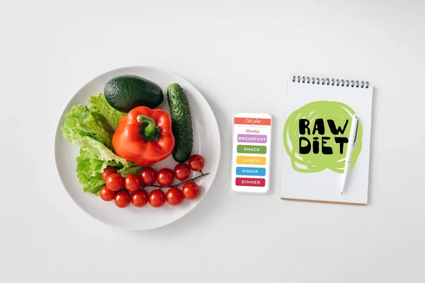 Вид сверху свежих овощей и авокадо на тарелке, смартфоне и ноутбуке на белом фоне — стоковое фото