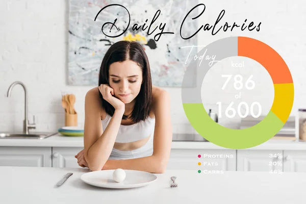 Pensive sportswoman looking at egg on kitchen table near daily calories illustration — Stock Photo