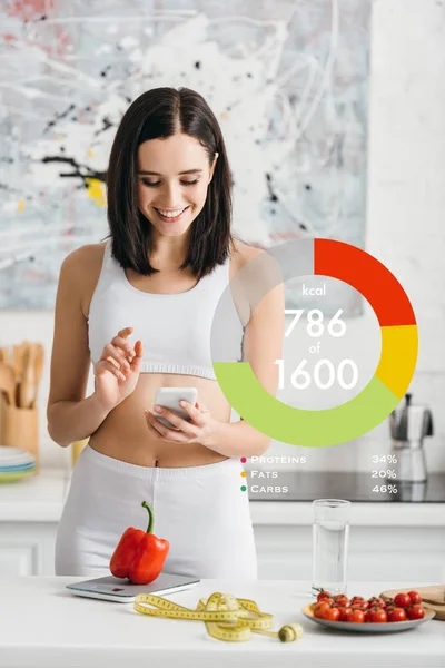 Smiling sportswoman using smartphone near measuring tape, vegetables and scales on kitchen table, calorie counting illustration — Stock Photo