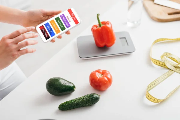 Vista recortada de niña sosteniendo teléfono inteligente con aplicación de plan de dieta diaria cerca de verduras, básculas y cinta métrica en la mesa de la cocina - foto de stock