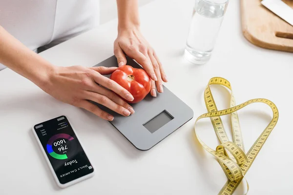 Vista cortada da mulher colocando tomate em escalas perto do smartphone com aplicativo de contagem de calorias e fita métrica na mesa da cozinha — Fotografia de Stock