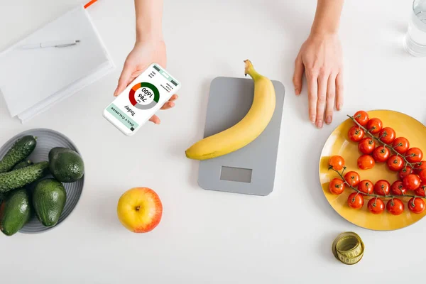 Vue du dessus de la fille tenant smartphone avec application de comptage des calories tout en pesant la banane sur la table de cuisine près des légumes frais — Photo de stock