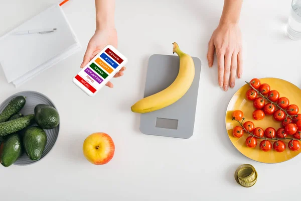 Top view of girl holding smartphone with diet plan while weighing banana on kitchen table with vegetables — Stock Photo