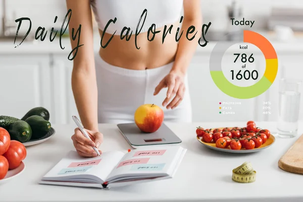 Vista recortada de la deportista en forma escribiendo calorías mientras pesa manzana en la mesa de la cocina, ilustración de calorías diarias - foto de stock