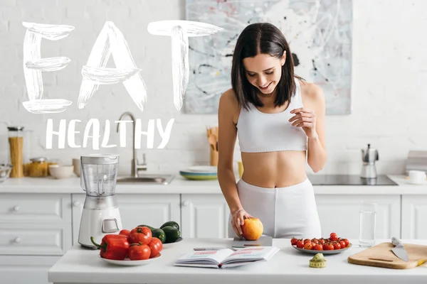 Deportista sonriente pesando manzana cerca del cuaderno y cinta métrica en la mesa de la cocina, comer ilustración saludable - foto de stock