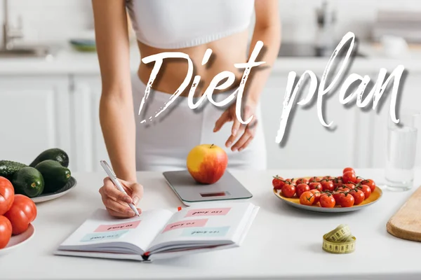 Vista recortada de la deportista en forma escribiendo calorías mientras pesa manzana en la mesa de la cocina, ilustración del plan de dieta - foto de stock