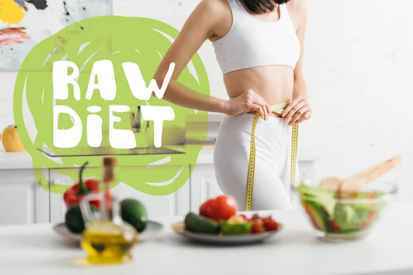 Selective focus of slim woman measuring waist with tape near fresh vegetables and salad on table, raw diet illustration — Stock Photo