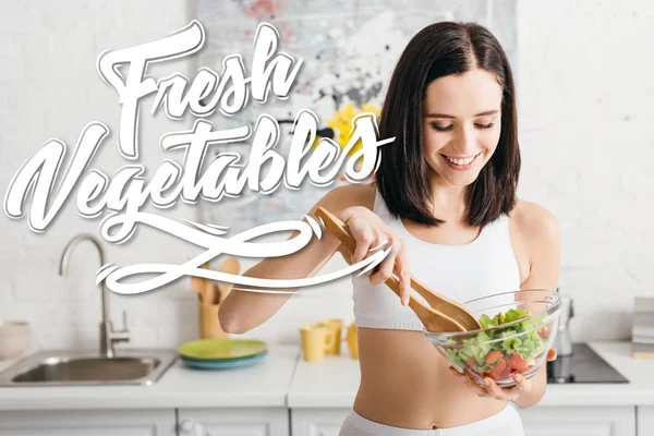 Attractive fit sportswoman smiling while mixing salad on kitchen, fresh vegetables illustration — Stock Photo