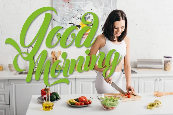 Mujer atractiva sonriendo mientras corta verduras orgánicas cerca del tazón con ensalada y cinta métrica en la mesa de la cocina, ilustración de buenos días - foto de stock