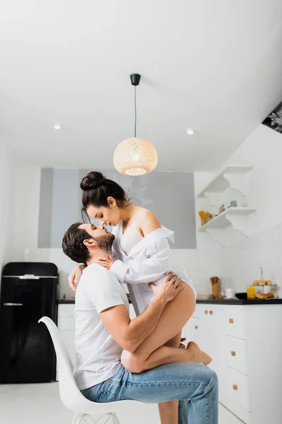 Vista laterale della ragazza sexy in reggiseno e camicia baciare fidanzato sulla sedia in cucina — Foto stock