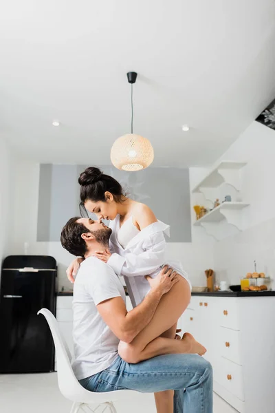 Vista lateral de chica sexy en sujetador y camisa besar novio en silla en la cocina - foto de stock