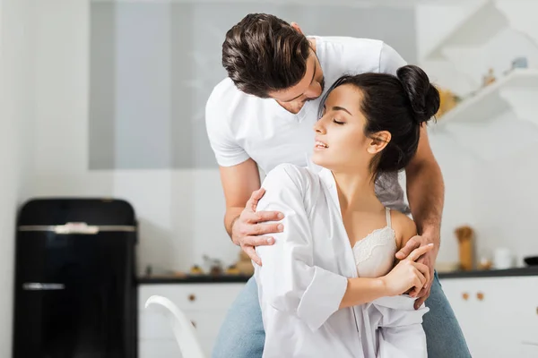 Man hugging attractive girlfriend in shirt and bra in kitchen — Stock Photo