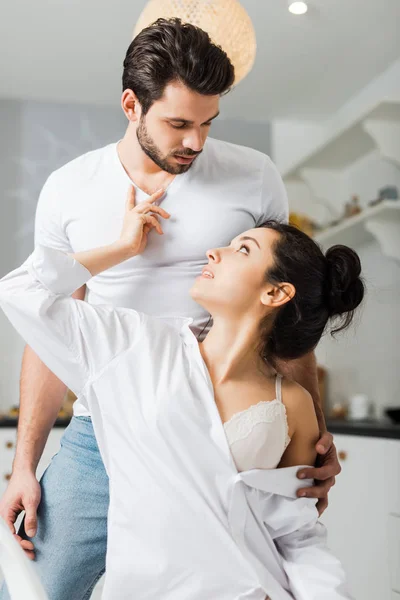 Sensual woman in shirt and bra touching chest of handsome boyfriend in kitchen — Stock Photo