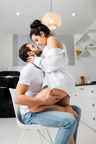 Vista lateral del hombre guapo tocando la cadera de novia sexy en sujetador y camisa en silla en cocina - foto de stock