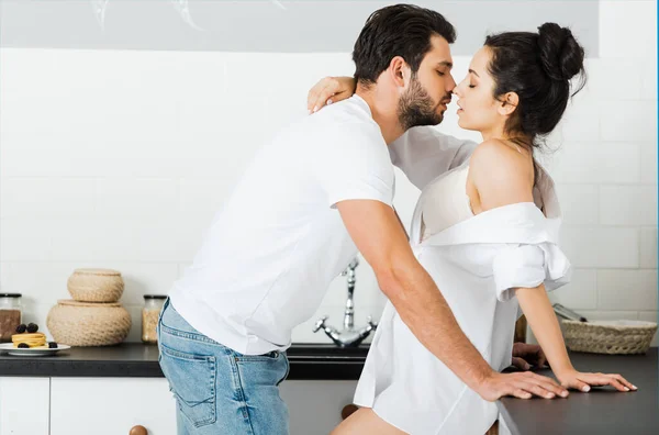 Vista laterale di uomo baciare donna sensuale in reggiseno e camicia in cucina — Foto stock