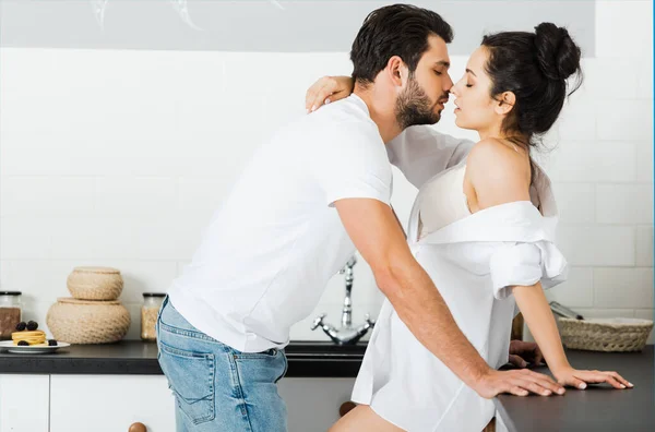 Side view of man kissing sensual woman in bra and shirt in kitchen — Stock Photo