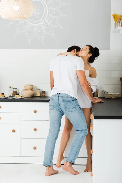 Мужчина целует в шею красивую женщину в лифчике и рубашку на кухне — стоковое фото