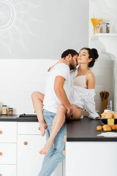 Hombre guapo besándose en el cuello y abrazando a novia seductora en sujetador y camisa en encimera de cocina - foto de stock