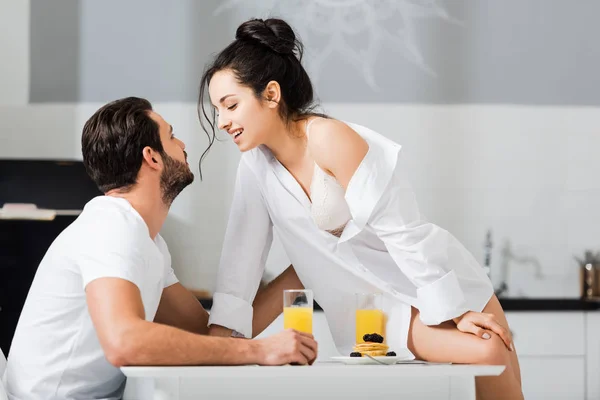 Vista lateral de la mujer sexy en camisa y sujetador sonriendo al novio mientras está sentado en la mesa de la cocina - foto de stock