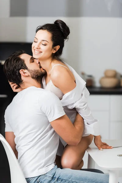 Bello uomo baciare donna sorridente in reggiseno e camicia in cucina — Foto stock