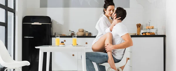 Panoramic shot of sexy woman kissing boyfriend on chair during breakfast in kitchen — Stock Photo