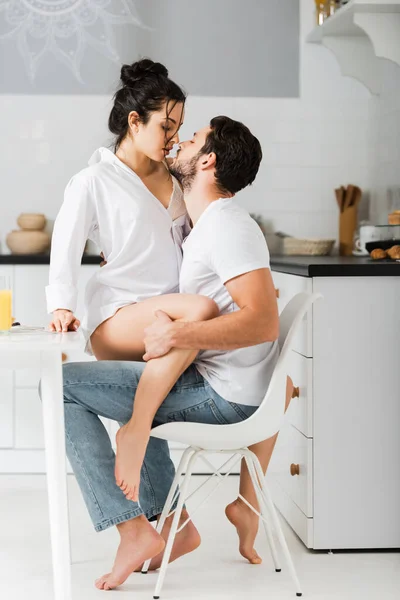 Vista lateral de mujer atractiva besando novio guapo en silla en la cocina - foto de stock