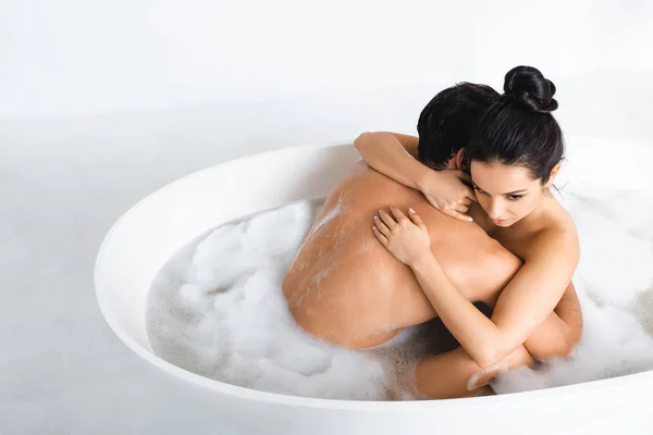 Hermosa mujer desnuda abrazando novio en la bañera con espuma en la superficie gris sobre fondo blanco - foto de stock