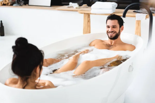 Selective focus of handsome man smiling at girlfriend in bathtub with foam — Stock Photo