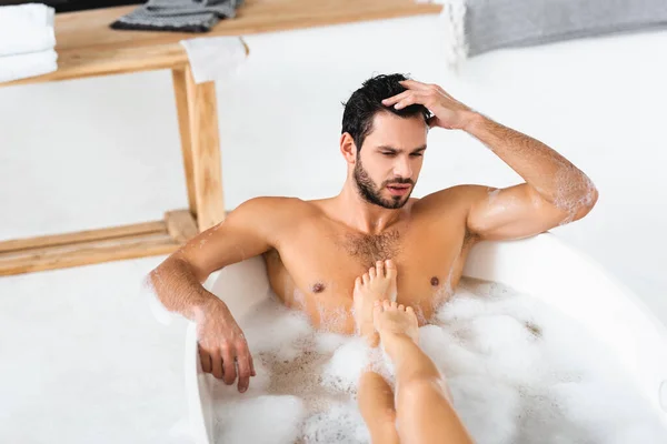 Vista de ángulo alto de los pies femeninos en el pecho del hombre guapo en la bañera con espuma - foto de stock