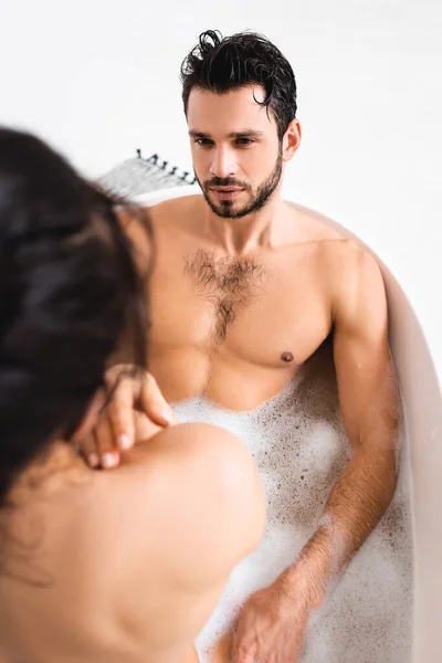 Selective focus of handsome muscular man looking at naked girlfriend in bathtub on white background — Stockfoto