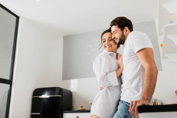 Vista lateral de homem bonito abraçando namorada sorridente na camisa perto da bancada da cozinha — Fotografia de Stock