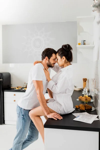 Vista lateral da mulher sensual abraçando namorado bonito enquanto sentado na bancada da cozinha — Fotografia de Stock