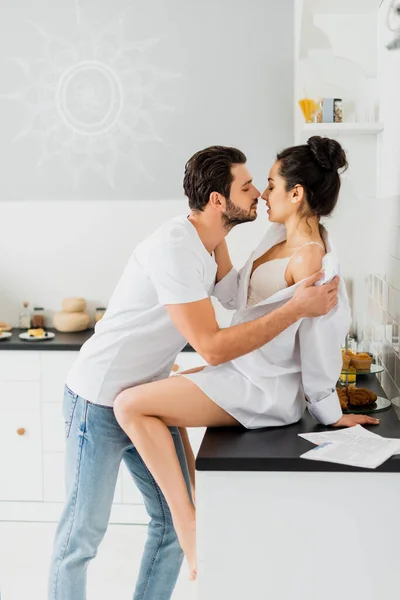 Perfil del hombre guapo quitándose la camisa de novia sexy en la encimera de la cocina - foto de stock