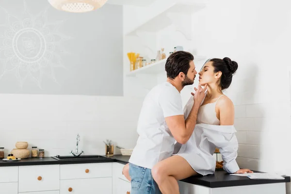 Vista lateral del hombre guapo besando novia sexy en sujetador y camisa en encimera de cocina - foto de stock