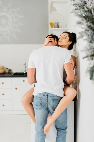 Vista trasera del hombre abrazando y besando sensual novia en encimera de la cocina - foto de stock