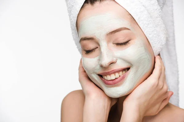 Chica sonriente con mascarilla facial nutritiva y toalla en la cabeza tocando la cara con los ojos cerrados aislados en blanco - foto de stock