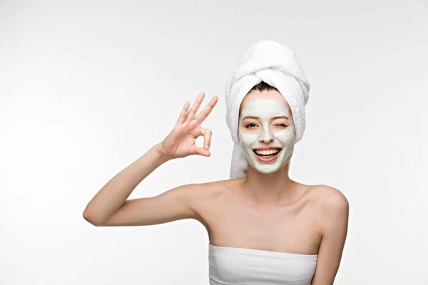 Fille heureuse avec masque facial nourrissant et serviette sur la tête montrant geste correct et clin d'oeil isolé sur blanc — Photo de stock