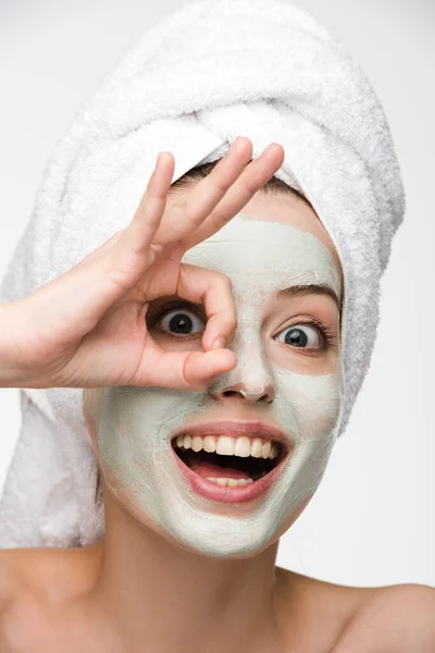 Chica alegre con mascarilla facial nutritiva y toalla en la cabeza mostrando gesto bien cerca de ojo aislado en blanco - foto de stock