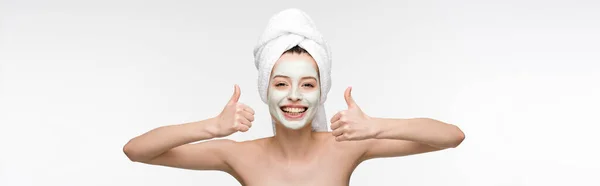 Plano panorámico de chica feliz con mascarilla facial nutritiva y toalla en la cabeza mostrando los pulgares hacia arriba aislado en blanco - foto de stock