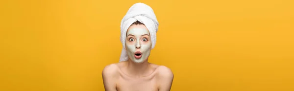 Panoramic shot of shocked girl with nourishing facial mask and towel on head looking at camera isolated on yellow — Stock Photo