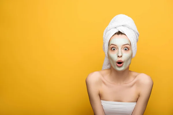 Шокированная девушка с питательной маской для лица и полотенцем на голове, смотрящая на камеру на желтом фоне — стоковое фото