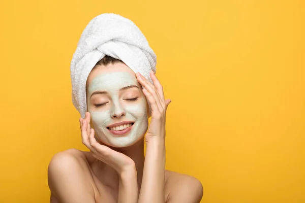 Menina feliz com máscara facial nutritiva e toalha na cabeça tocando o rosto com os olhos fechados isolados no amarelo — Fotografia de Stock