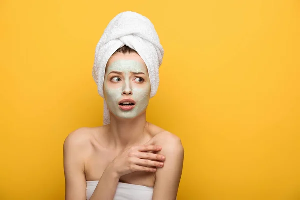 Ragazza spaventata con maschera nutriente viso e asciugamano sulla testa guardando lontano isolato sul giallo — Foto stock