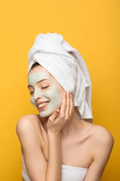 Chica feliz con mascarilla facial nutritiva y toalla en la cabeza tocando la cabeza con los ojos cerrados aislados en amarillo - foto de stock