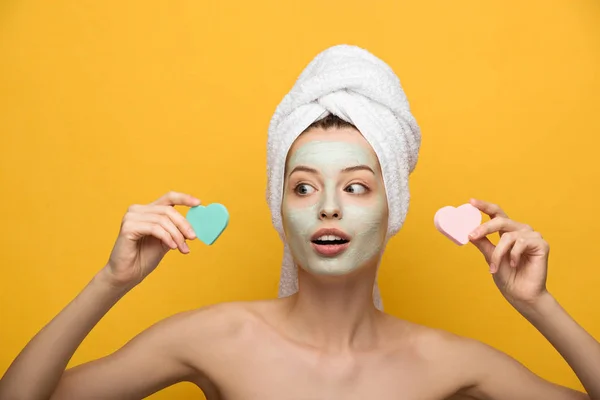 Surprised girl with nourishing facial mask holding heart-shaped cosmetic sponges and looking away on yellow background — Stock Photo