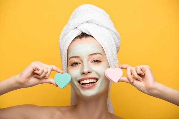 Menina sorridente com máscara nutritiva no rosto segurando esponjas cosméticas em forma de coração no fundo amarelo — Fotografia de Stock