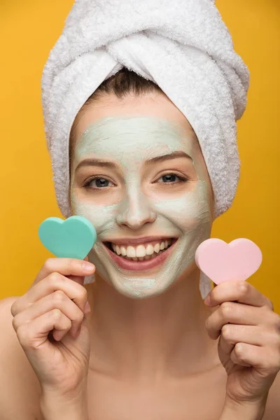 Cheerful girl with nourishing mask on face holding heart-shaped cosmetic sponges isolated on yellow — Stock Photo